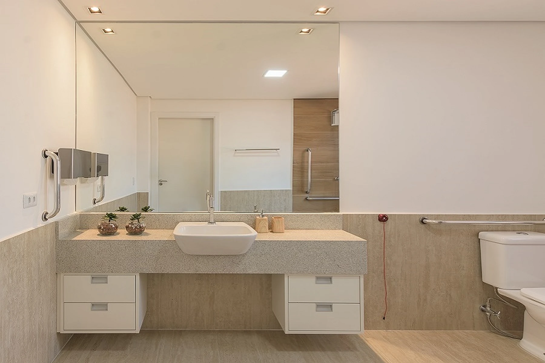 Banheiro moderno e acessível com barras de apoio em aço escovado ao lado do vaso sanitário e dentro do box de vidro. O espaço possui piso antiderrapante, um balcão com cuba esculpida e espelho iluminado.