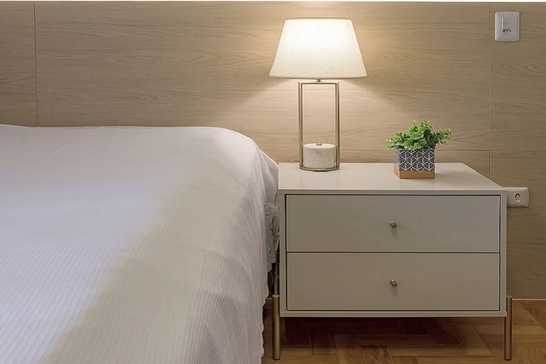 Um quarto de casa de repouso com cama de solteiro, criado-mudo e luminária, mostrando um ambiente limpo e acolhedor.