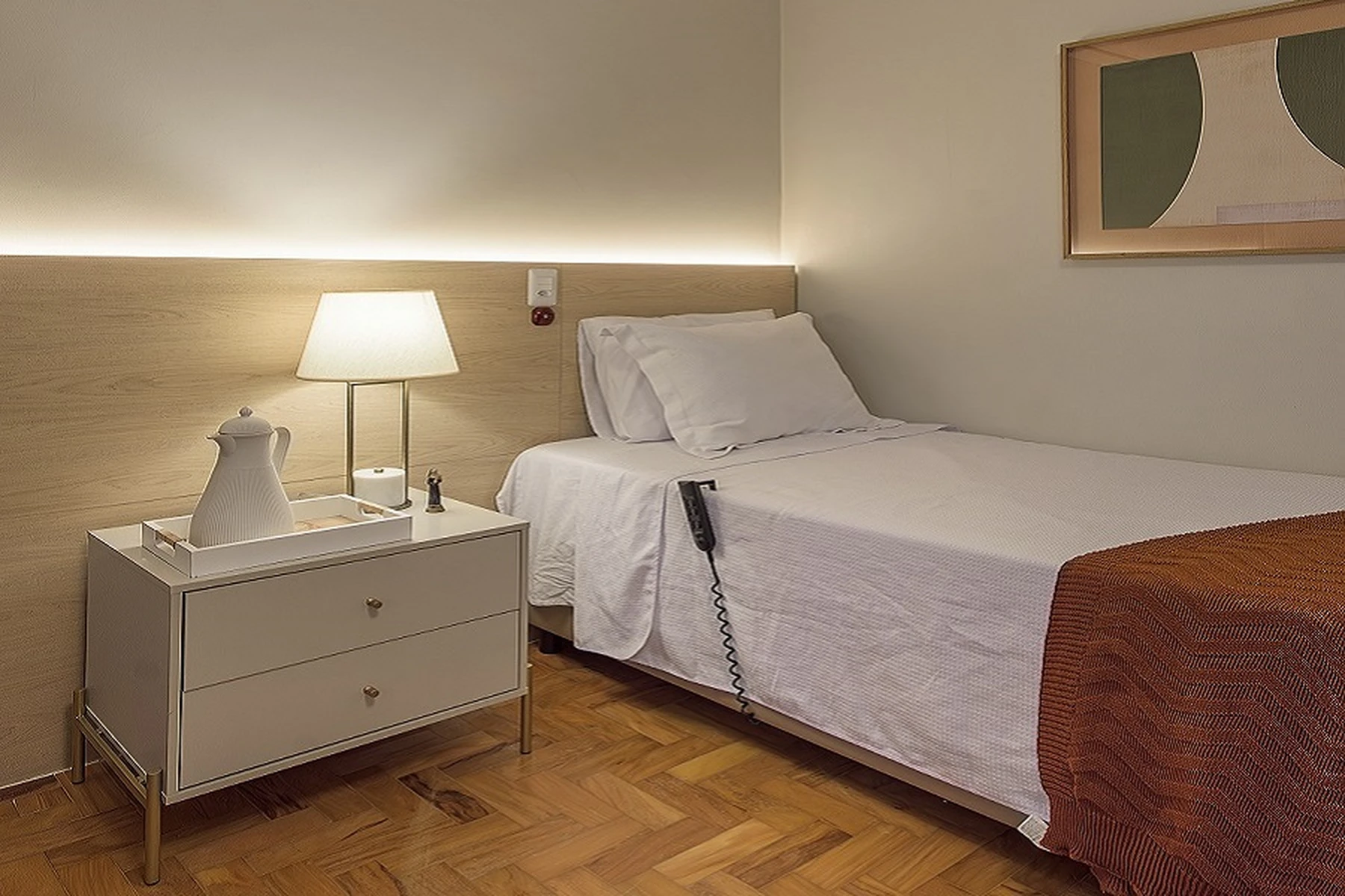 Um quarto de casa de repouso com cama de solteiro, criado-mudo e luminária, mostrando um ambiente limpo e acolhedor.
